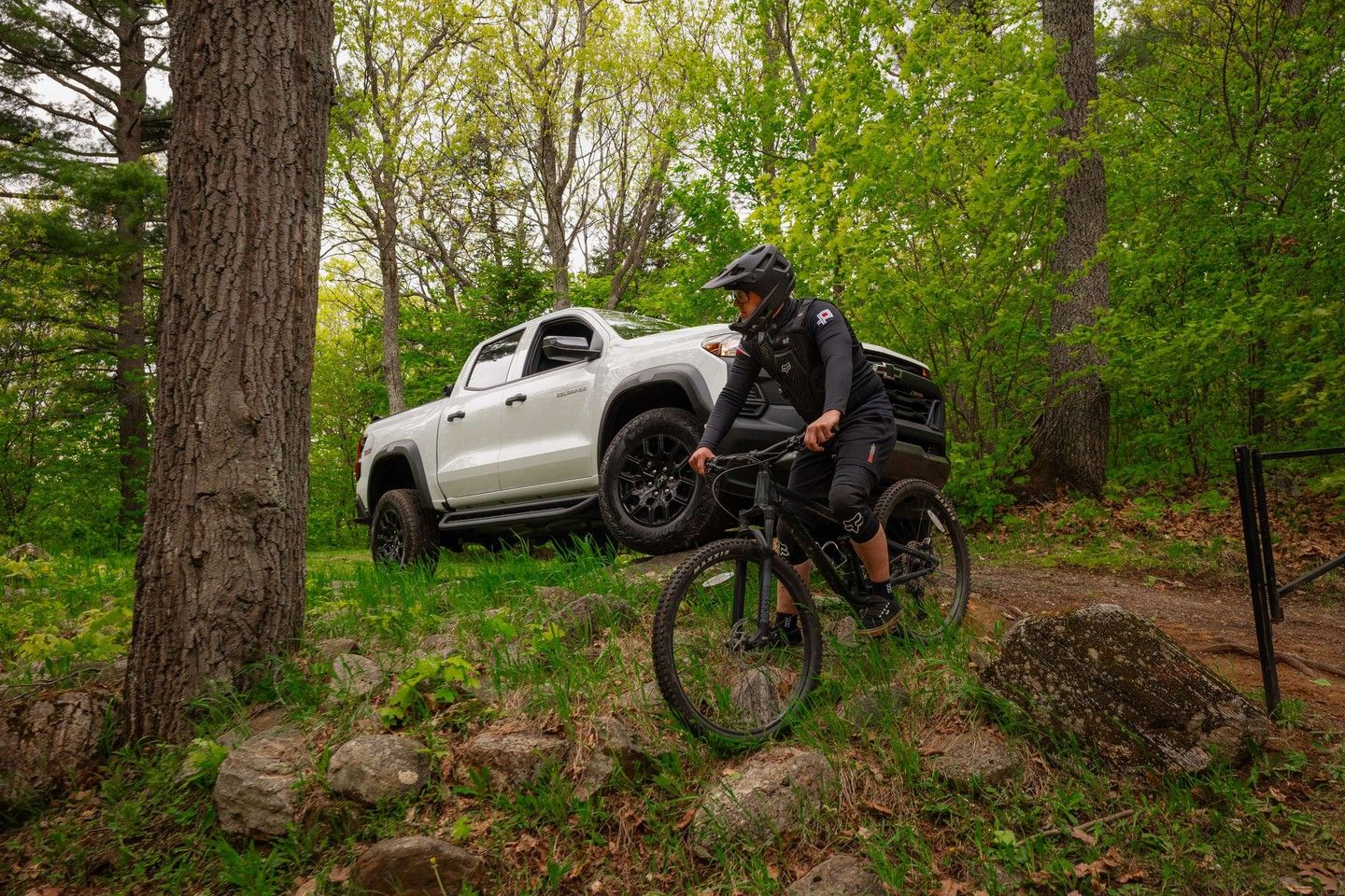A cyclist in off-road gear races down a forest trail, while a 2026 Chevrolet Colorado waits in the background—a perfect illustration of the truck as an adventure companion.