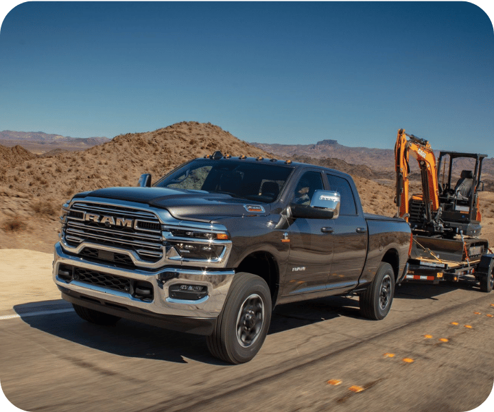 3/4 front and side view of the 2026 RAM 2500 on the road, towing a trailer and a backhoe