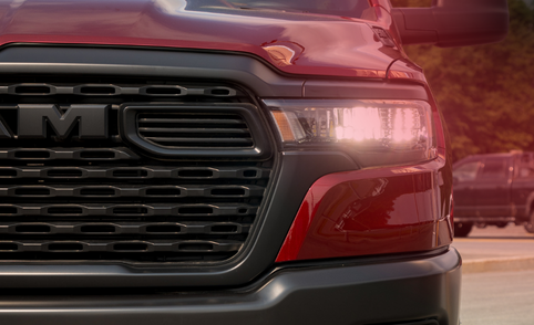 Close-up of the front end of a red 2026 RAM 1500 with its grille and headlight illuminated.