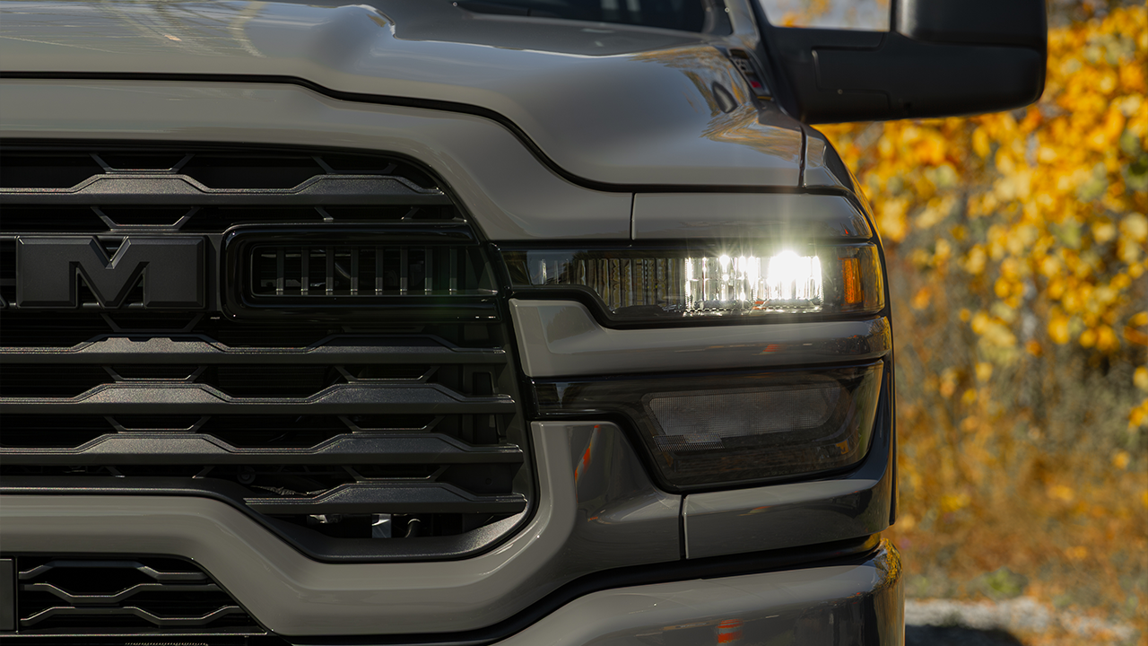 A close-up of the grille and LED headlight of a gray Ram pickup truck, with the lights on, against a backdrop of golden autumn foliage.