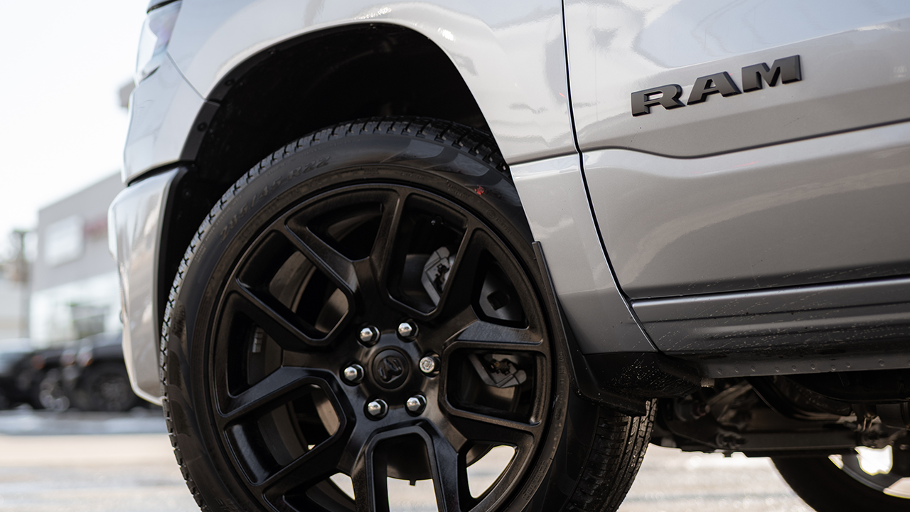 A close-up of the black-rimmed wheel and front fender of a silver-gray Ram pickup truck on a dealership lot.