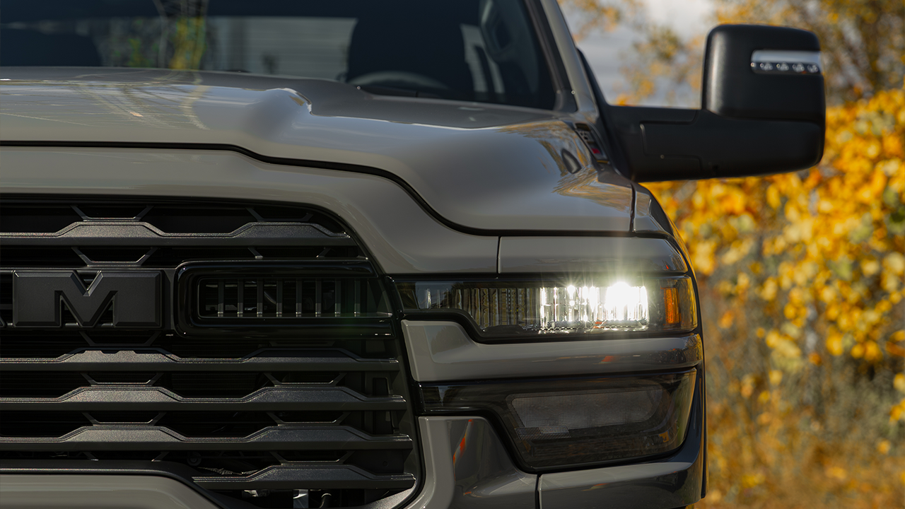 Close-up of the grille and the illuminated LED headlight of a gray Ram 2500, with a side mirror in the foreground and golden autumn foliage in the background.