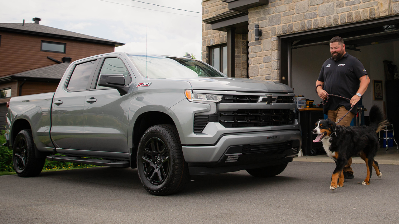 Vue 3/4 avant et lat&eacute;rale du Chevrolet Silverado 2026 stationn&eacute; devant un garage avec un sp&eacute;cialiste Paill&eacute; sortant son chien