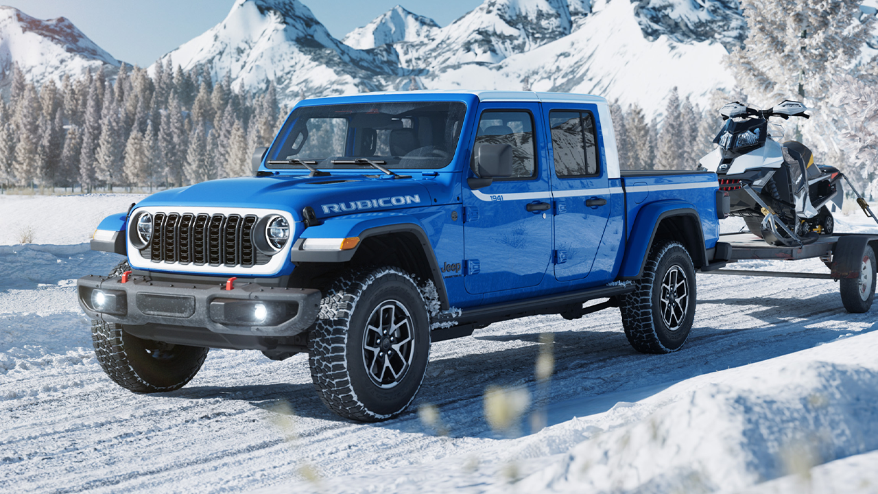 A 3/4 front and side view of the 2026 Jeep Gladiator on a snow-covered road, towing a trailer with a snowmobile.