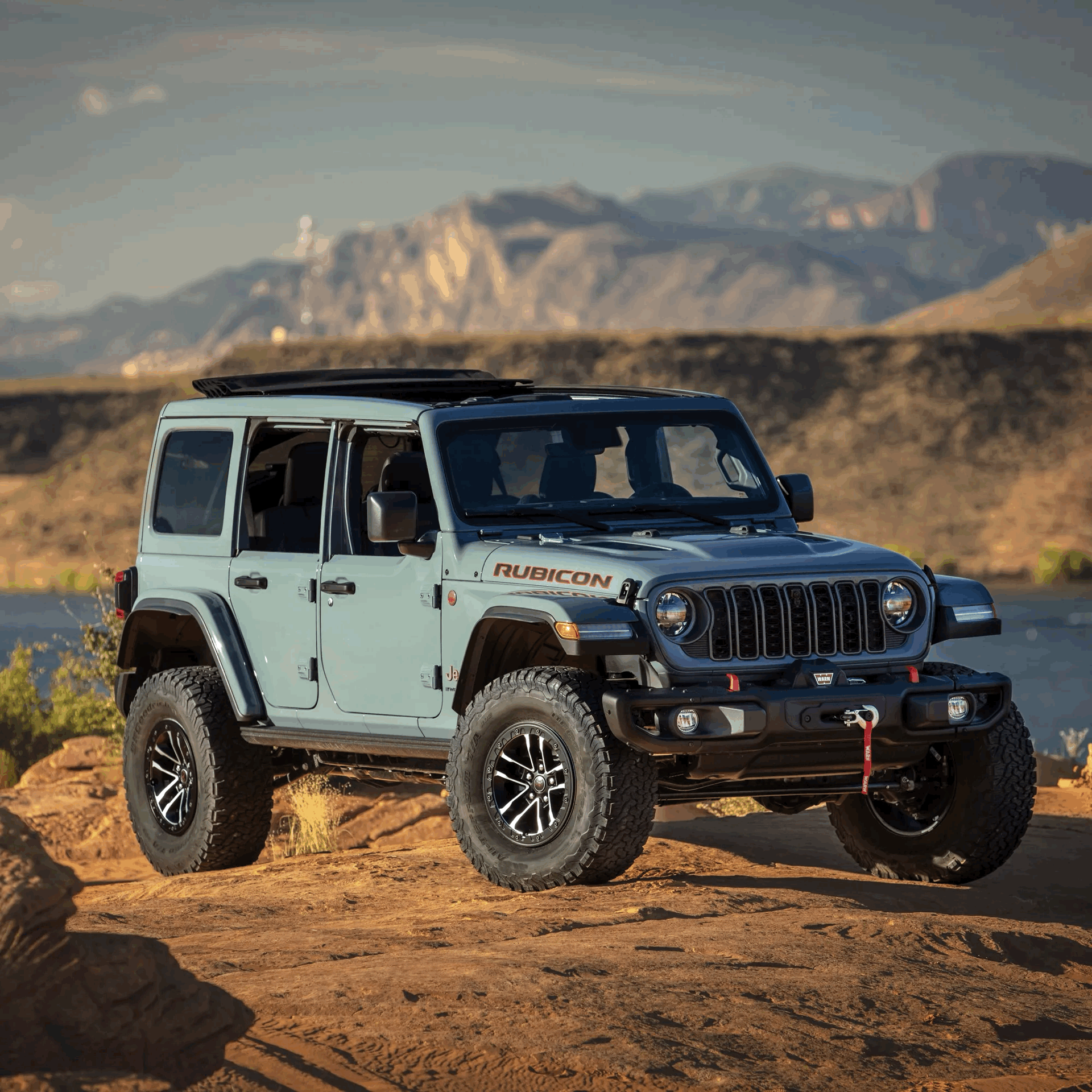 Grey-blue 4-door Jeep Wrangler Rubicon on rocky terrain with mountains in the background.
