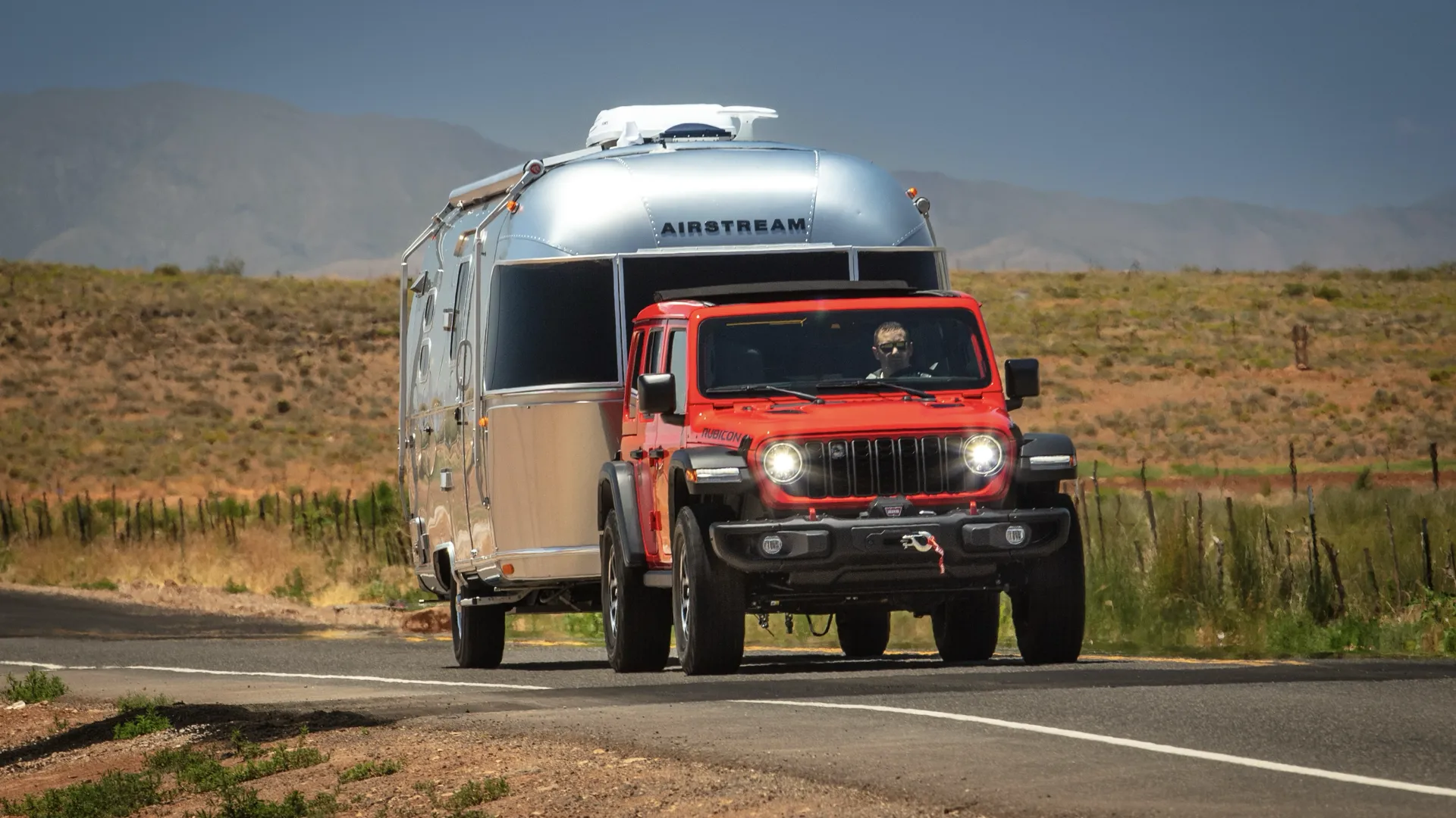 Red 4-door Jeep Wrangler towing an Airstream trailer on a country road.