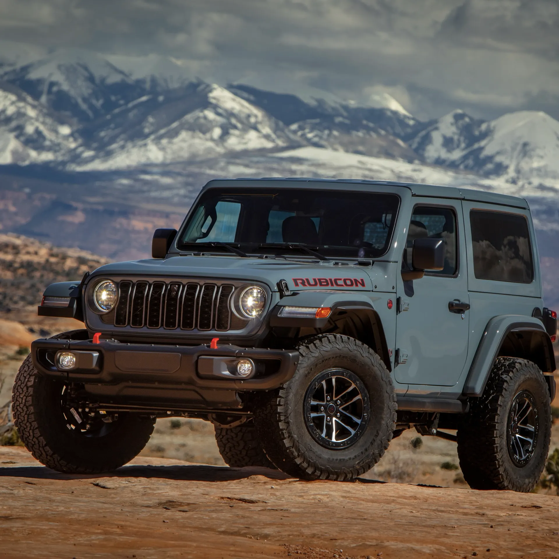 Grey-blue Jeep Wrangler Rubicon seen from the front in front of snow-capped mountains.