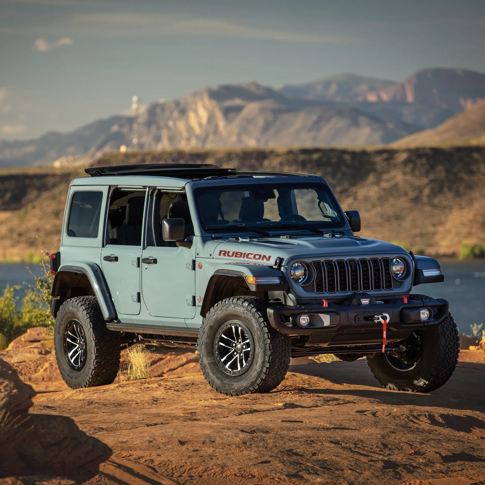Grey-blue 4-door Jeep Wrangler Rubicon with the top removed, on rocky terrain near a lake.