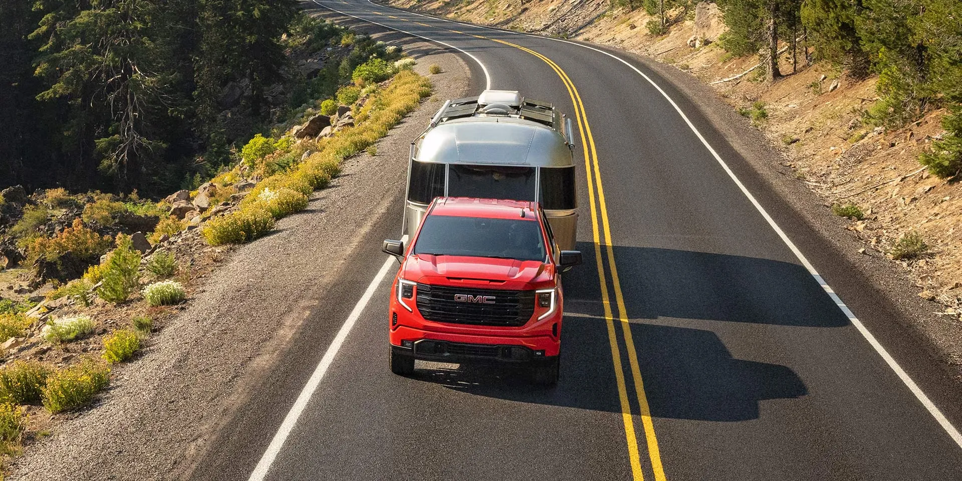 GMC Sierra 1500 2026 rouge remorquant une caravane sur route de montagne.