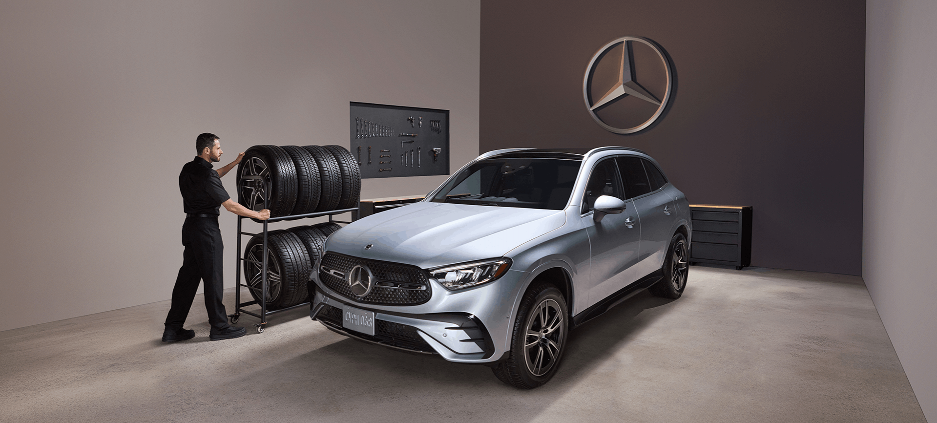 2026 Mercedes-Benz GLC - a silver 2026 Mercedes-Benz GLC SUV parked in a service bay next to a technician and a rack of tires - Mercedes-Benz Brampton - New car dealership in Brampton, Ontario