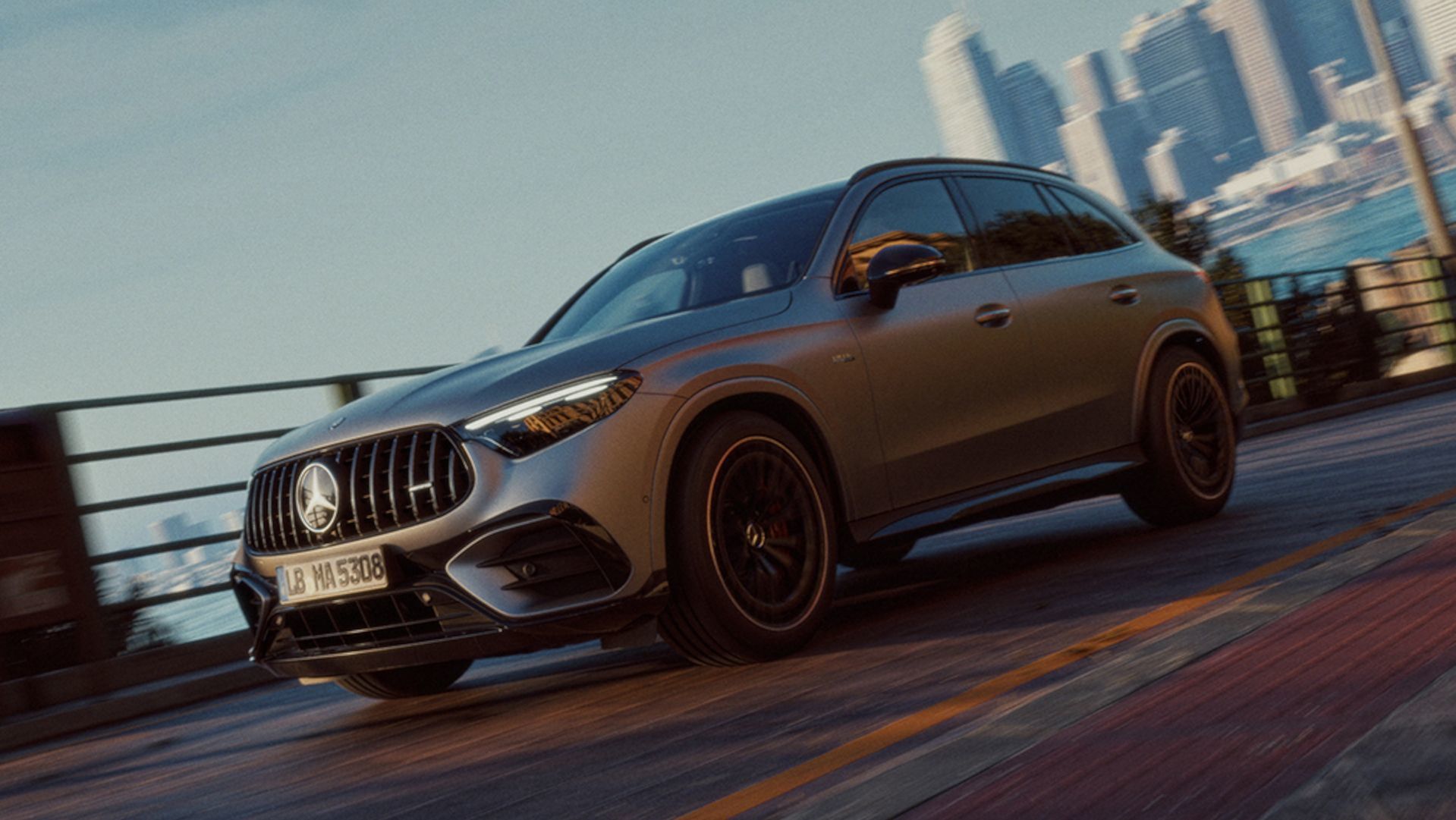 2026 Mercedes-AMG GLC - Action shot of a matte grey 2026 Mercedes-AMG GLC driving on a bridge with a city skyline in the background - Mercedes-Benz Brampton - New car dealership in Brampton, Ontario