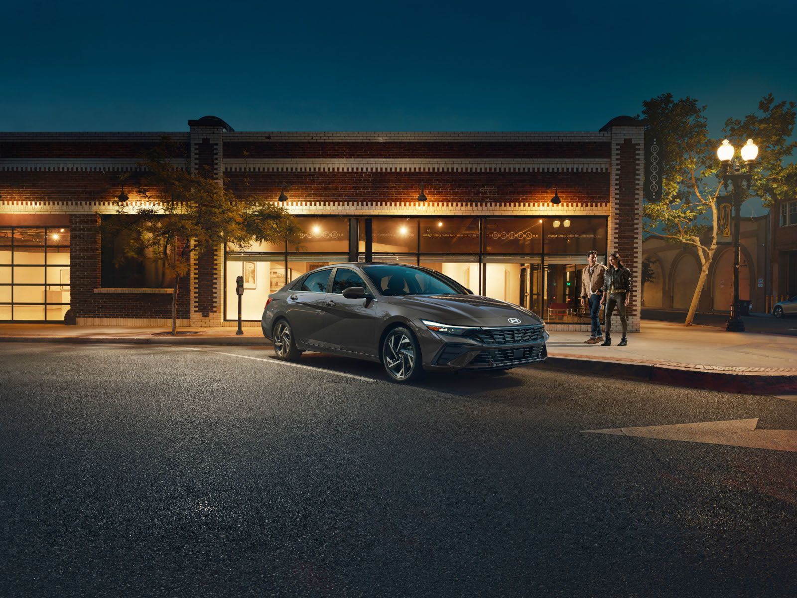 2026 Hyundai Elantra - A grey 2026 Hyundai Elantra sedan parked at night in an urban setting, with people walking in the background - Woodstock Hyundai - New car dealership in Woodstock, Ontario