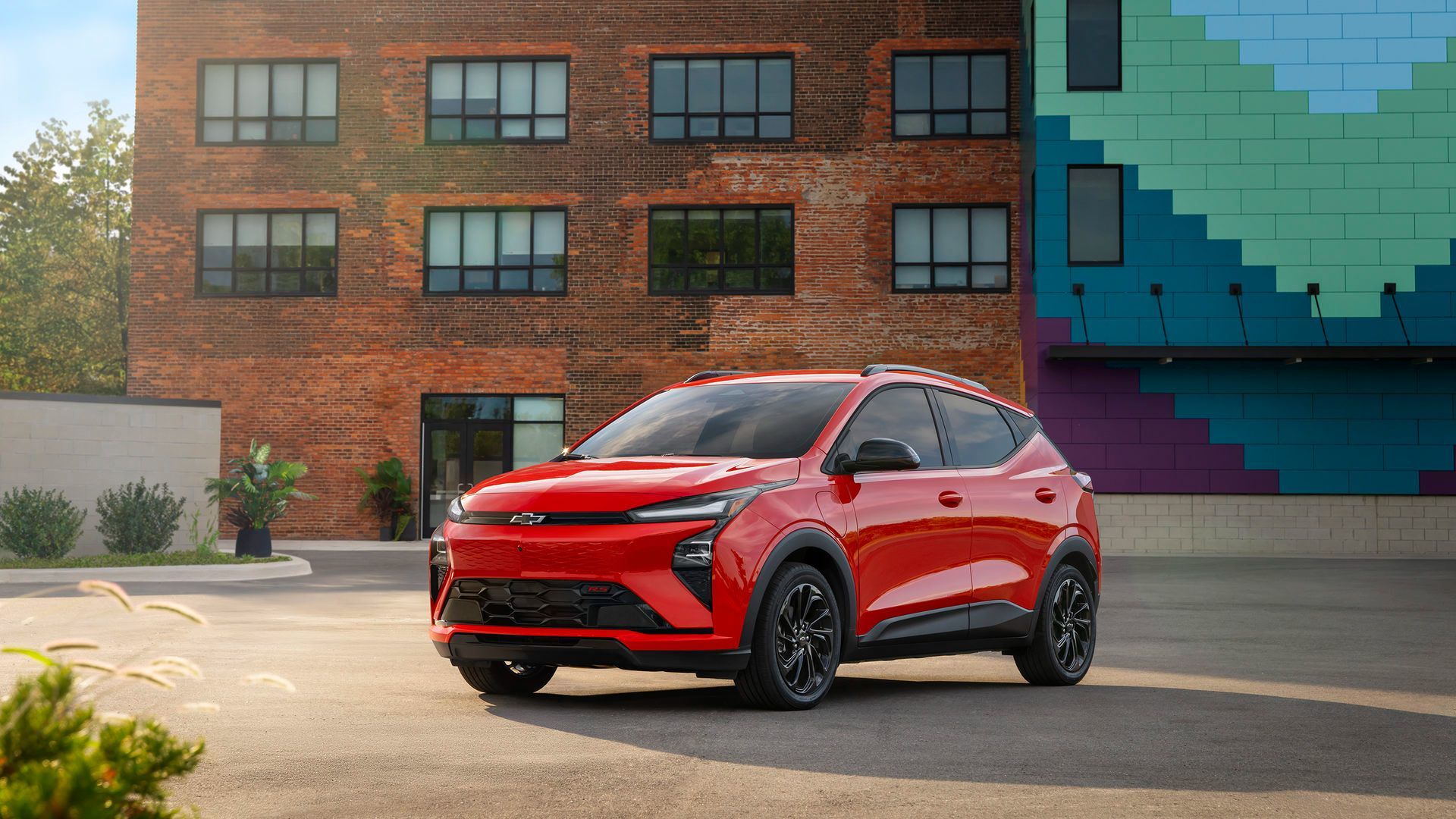 2027 Chevrolet Bolt - Front 3/4 view of a red Chevrolet Bolt parked in an urban parking lot in front of a brick building and a modern blue and green geometric wall - Applewood Chevrolet, Buick, GMC - New car dealership in Mississauga, Ontario