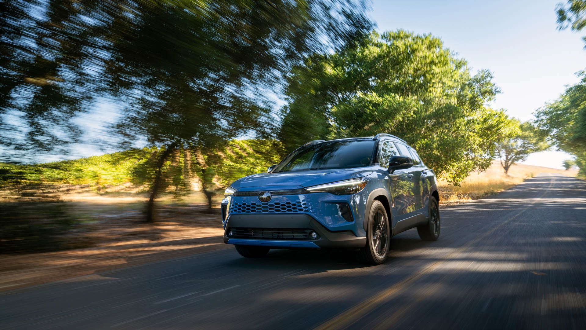 2026 Toyota Corolla Cross - a blue 2026 Toyota Corolla Cross driving on a tree-lined road - Oakville Toyota - Toyota car dealership in Oakville, Ontario