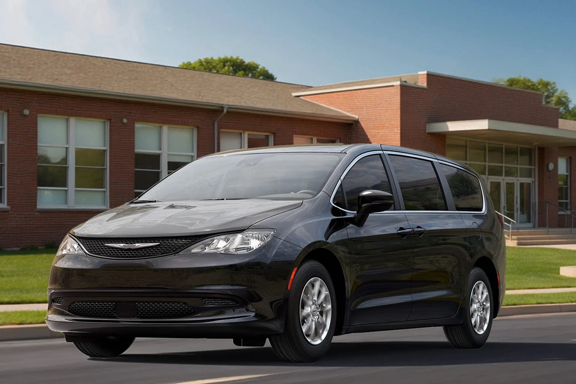 Black 2027 Chrysler Grand Caravan driving past a school, shown from the front three-quarter angle.