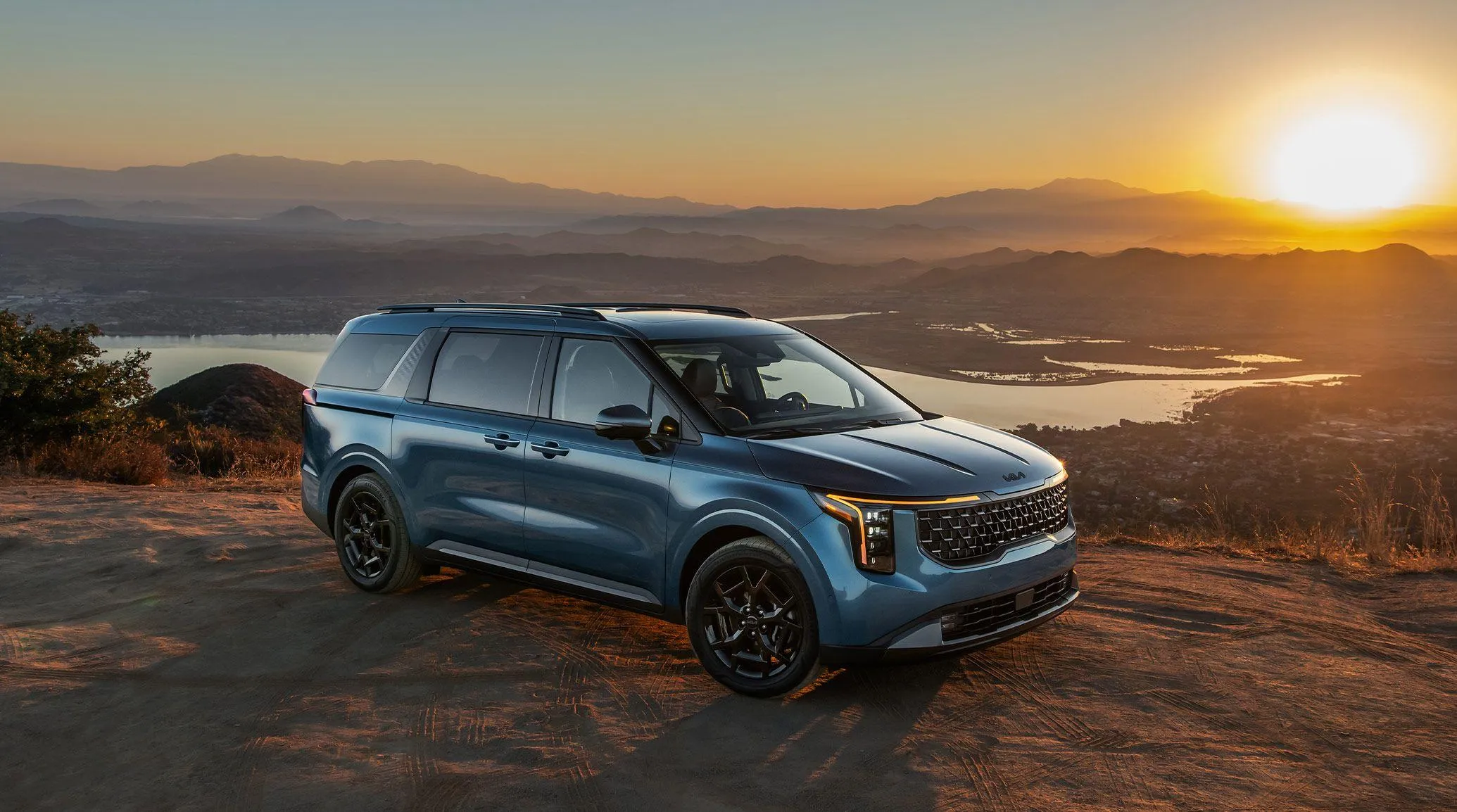Blue 2027 Kia Carnival parked overlooking a rocky landscape at sunset.