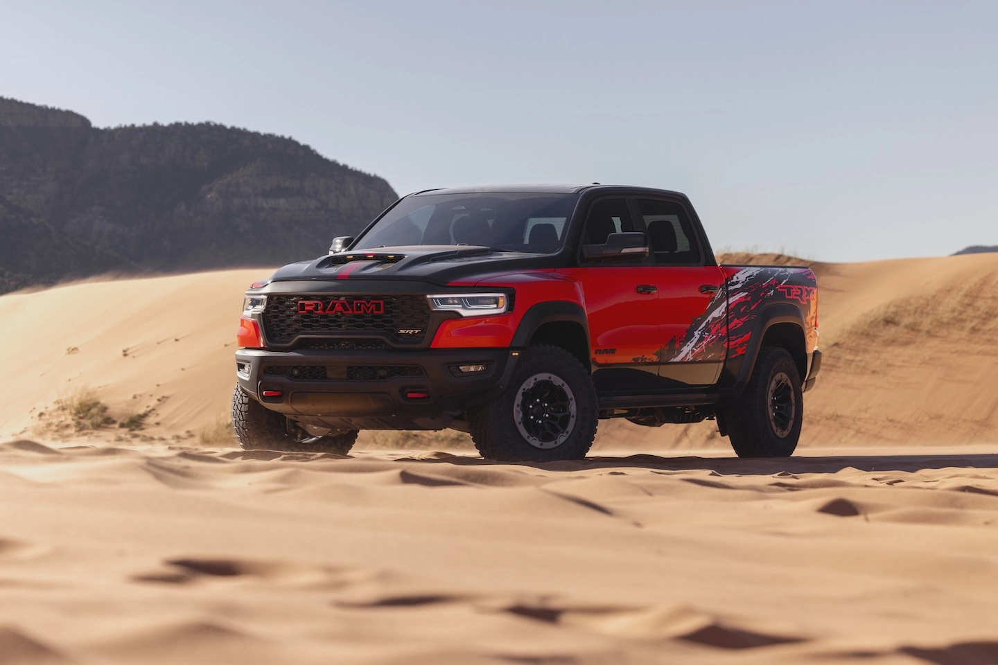 Red 2027 Ram 1500 SRT TRX with a black hood and body graphics parked on sand dunes.