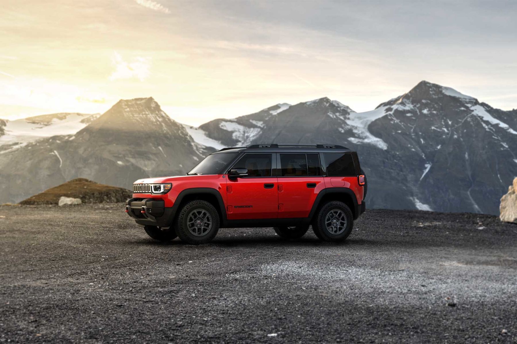 Jeep Recon 2026 rouge vu de profil stationn&eacute; sur un terrain rocheux en montagne au coucher du soleil.
