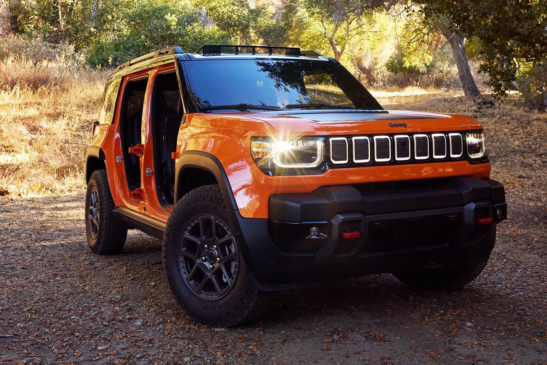 Jeep Recon 2026 orange avec portes retir&eacute;es stationn&eacute; en for&ecirc;t, vue trois quarts avant.
