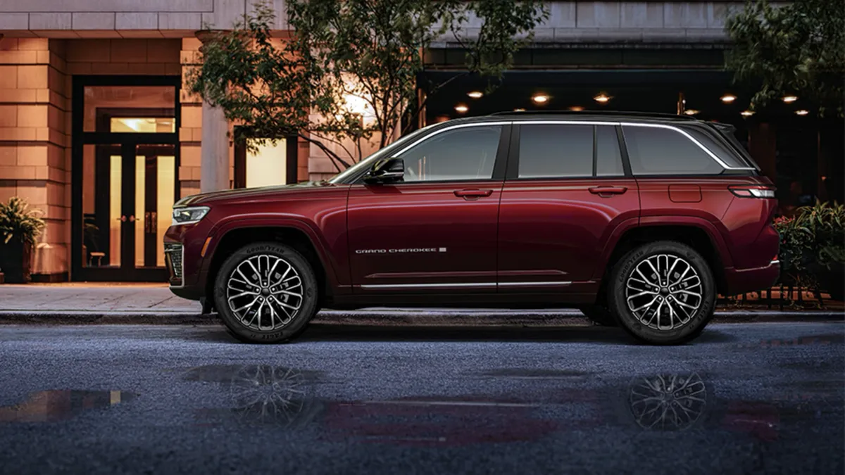 Jeep Grand Cherokee 2026 rouge bourgogne vu de profil devant une r&eacute;sidence moderne en milieu urbain.
