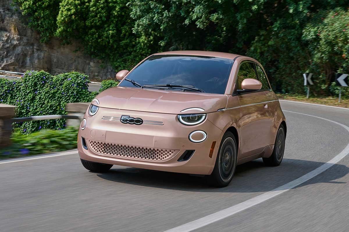 Beige 2025 Fiat 500e driving on a winding road surrounded by greenery.