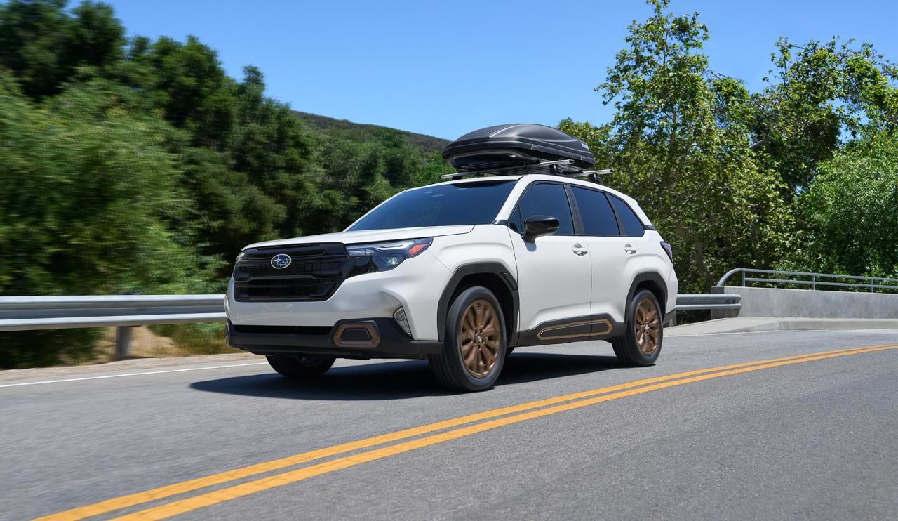 Subaru Forester 2026 blanc avec coffre de toit roulant sur une route boisée.