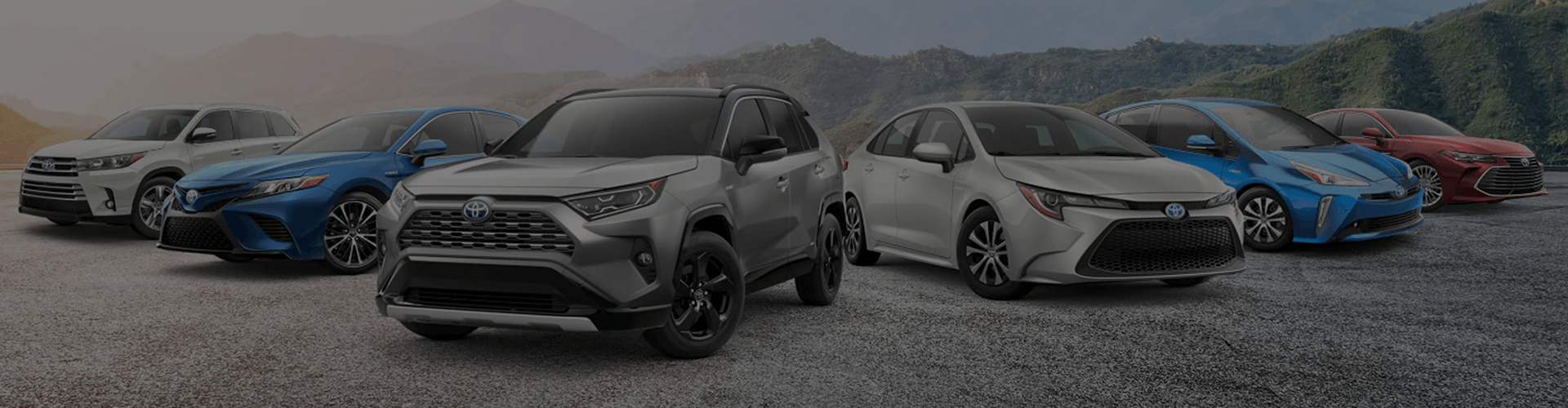 A line of six Toyota hybrid vehicles (SUVs and sedans) of various colors is parked on a gravel surface with a hazy mountainous background.