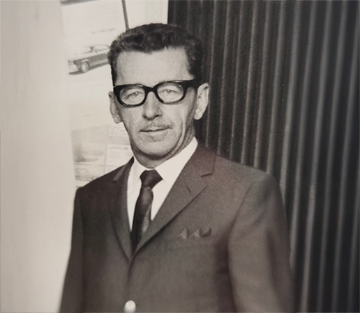 Black and white portrait of Mr. Ovila Arbour, founder of Arbour Automobiles, wearing dark-rimmed glasses, a suit, and a tie. In the background, an AMC (American Motor Company) car poster is visible, reflecting the company’s early years and heritage.
