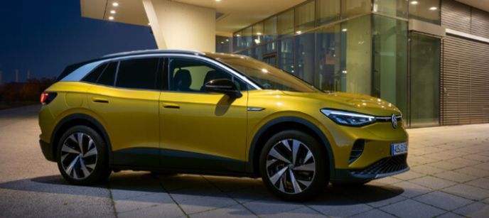 Vue latérale d'un VUS électrique Volkswagen ID.4 jaune métallisé stationné devant un bâtiment moderne vitré au crépuscule.