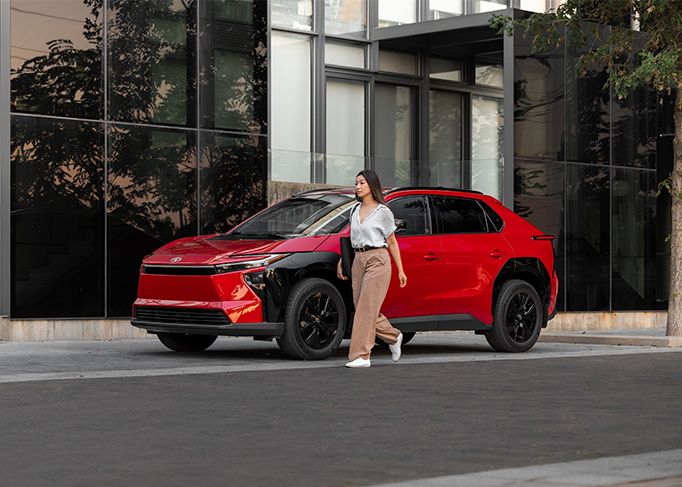 A red sedan is parked on a mountain road, with a large rocky cliff in the background.