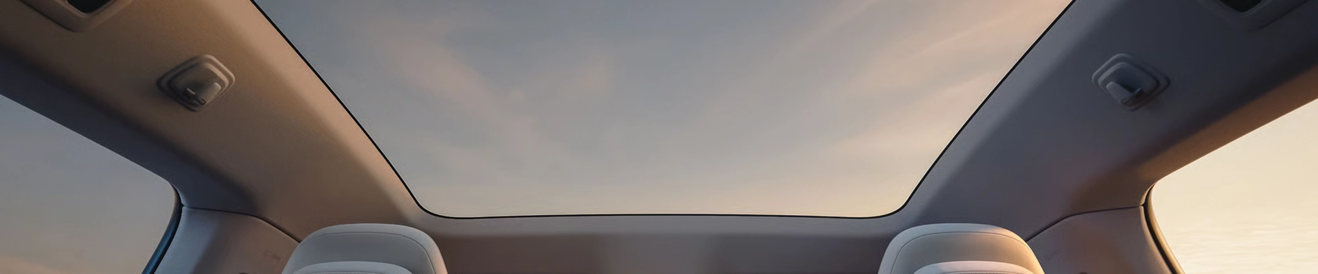 Interior view looking up through a large panoramic glass sunroof of a modern vehicle toward a soft sunset sky.