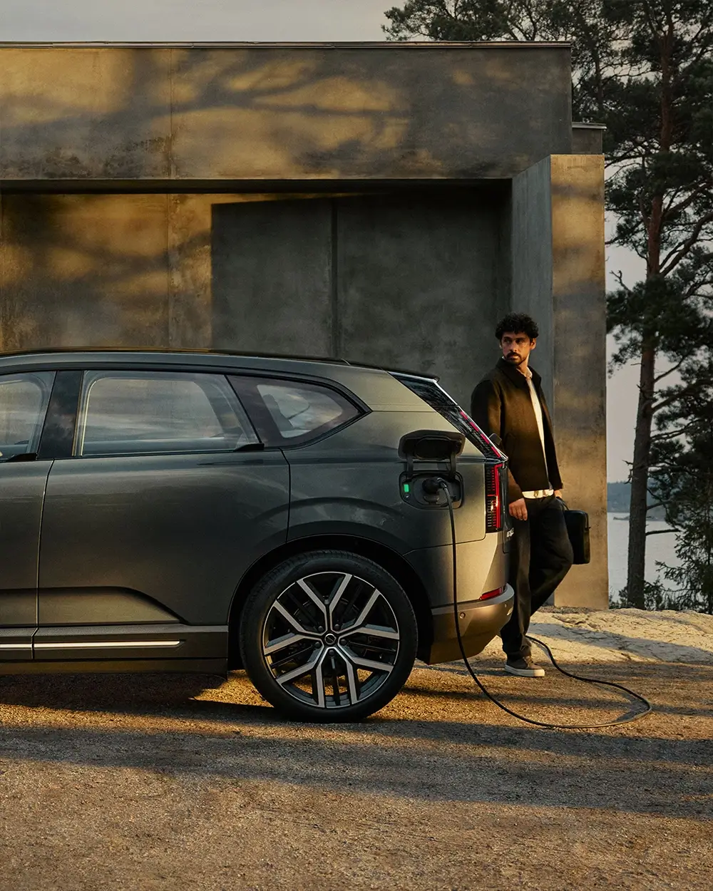 A man holding a briefcase leans against a dark gray Volvo electric SUV plugged into a charging cable outside a modern home.