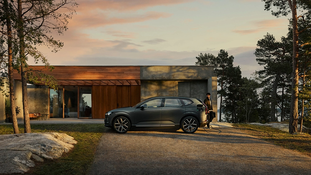 Profile view of a dark gray Volvo electric SUV plugged into a home charger outside a modern house with wood and concrete siding, framed by tall pine trees.