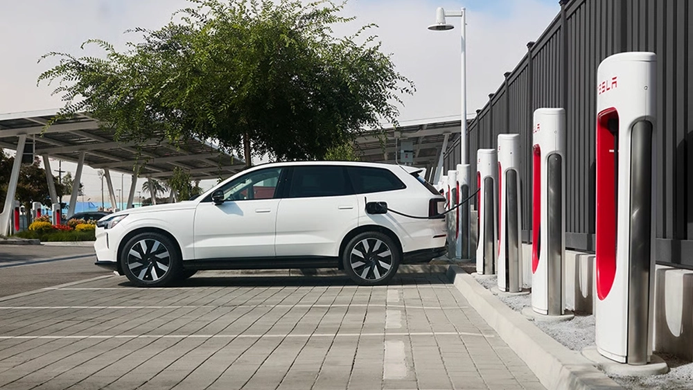 A white Volvo electric SUV parked at an outdoor charging plaza, plugged into a row of Tesla Superchargers.