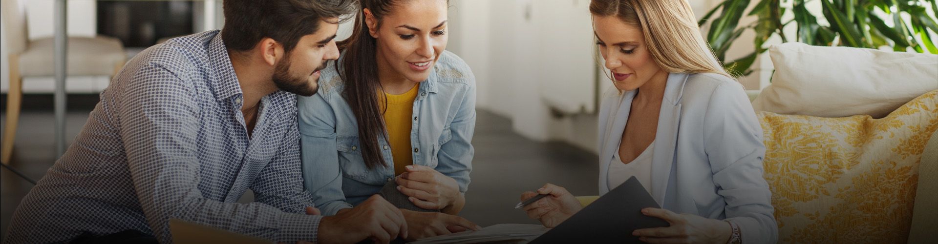 A couple meets with a financial advisor in a professional and casual setting. The advisor, a woman with long blonde hair, sits opposite the couple and points to a document. The couple, a man with a beard and a woman with long dark hair, looks at the document with interest.