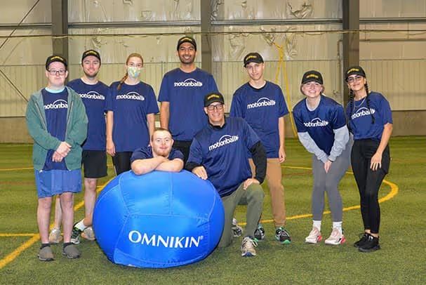 A group of nine people in matching blue "motionball" shirts and black caps posing in an indoor sports facility. In the foreground, a man leans on a large, bright blue "OMNIKIN" inflatable ball.