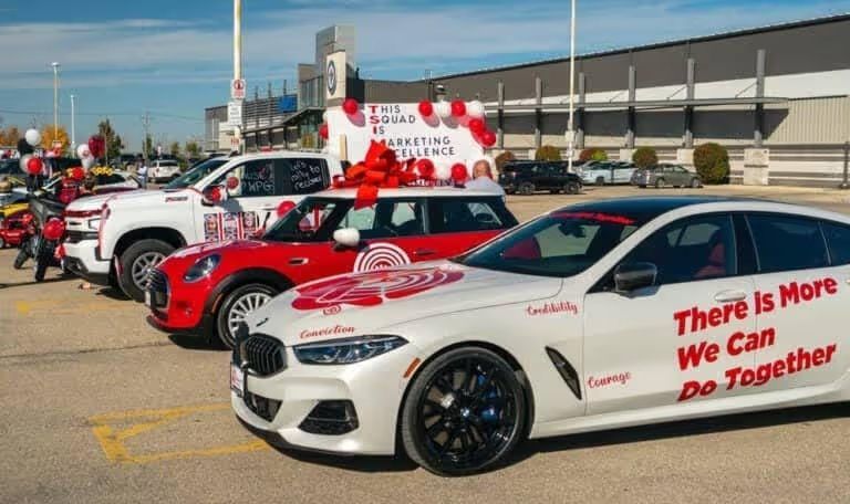 A lineup of several vehicles decorated for a corporate or charity event. The lead car is a white BMW featuring the text "There Is More We Can Do Together" and words like "Courage" and "Conviction" in red lettering.
