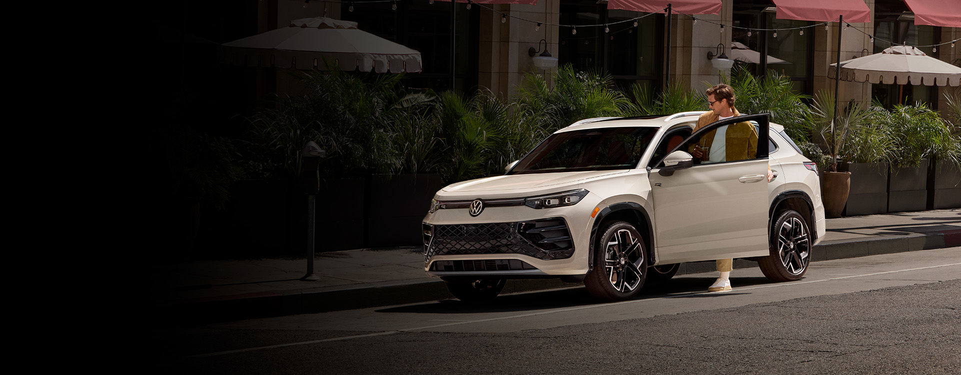  A light-colored Volkswagen Taos is parked on a city street with a person partially visible getting out of the driver's side door. The background shows building facades with awnings and potted plants.