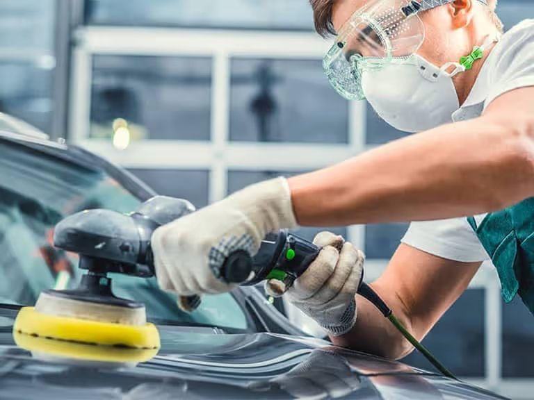 A close-up shot of an automotive technician at work. The individual is wearing clear safety goggles, a white respirator mask, and protective gloves. They are holding a professional dual-action orbital polisher with a yellow foam pad, applying it to the hood of a dark, glossy car. The background shows a bright, clean automotive shop environment with large windows.