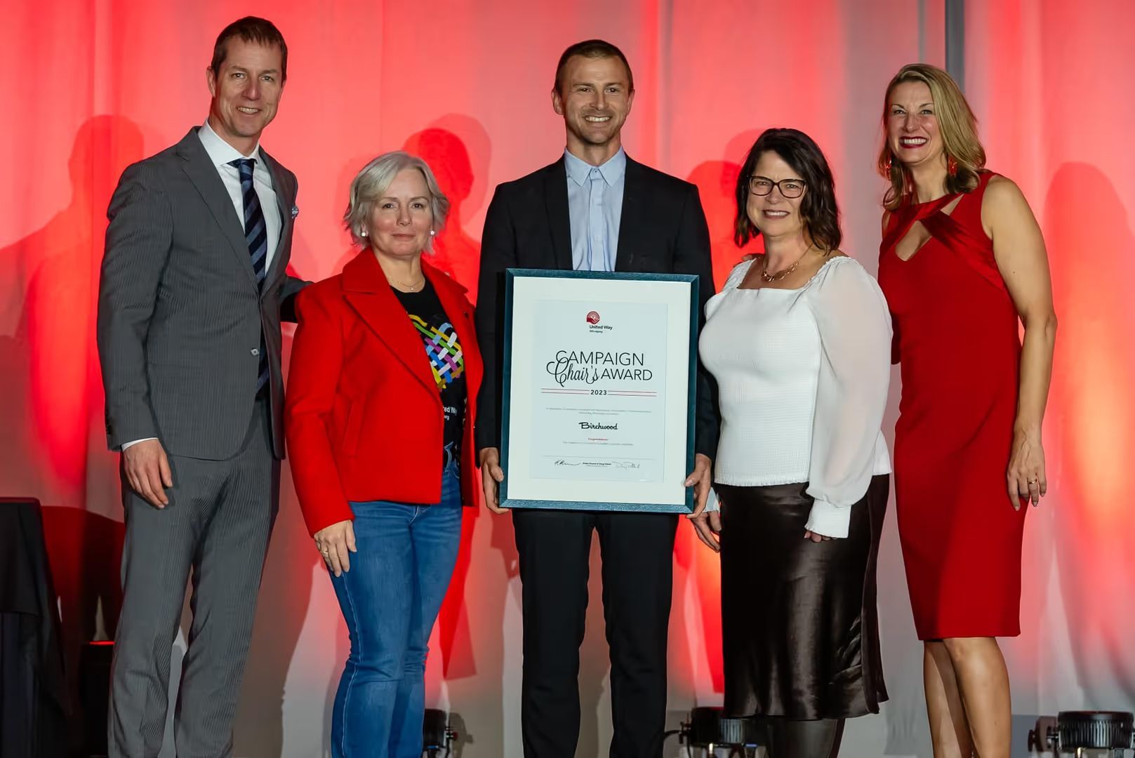 Five people standing on a stage in front of a red backdrop, holding a framed "2023 Campaign Chair’s Award" from United Way Winnipeg presented to Birchwood.