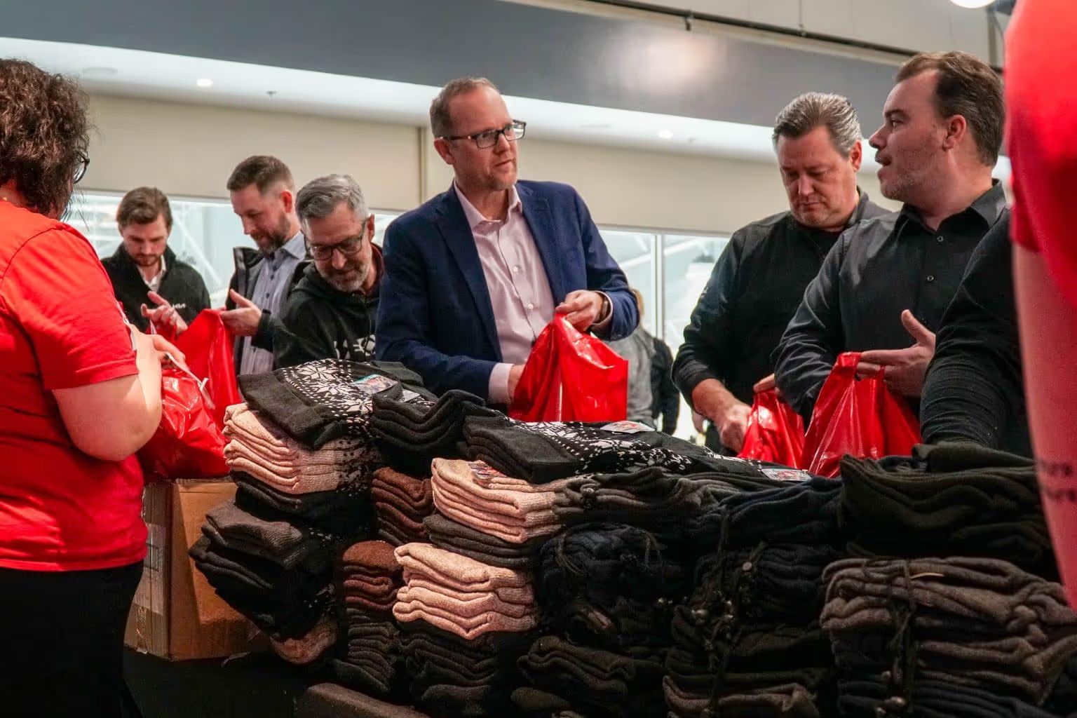 A group of people volunteering indoors, packing folded winter clothing into red plastic bags at a long table.