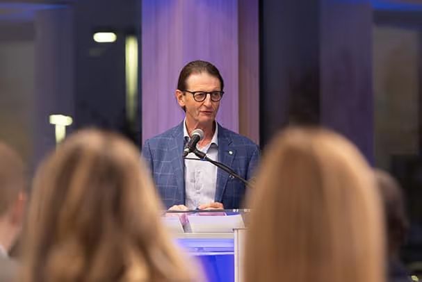 A man in a blue checkered blazer and glasses speaking at a microphone behind a podium, viewed from behind an audience.