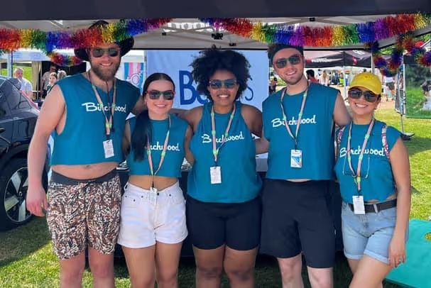 Five people wearing matching teal "Birchwood" tank tops and sunglasses standing together under a tent decorated with rainbow garland.