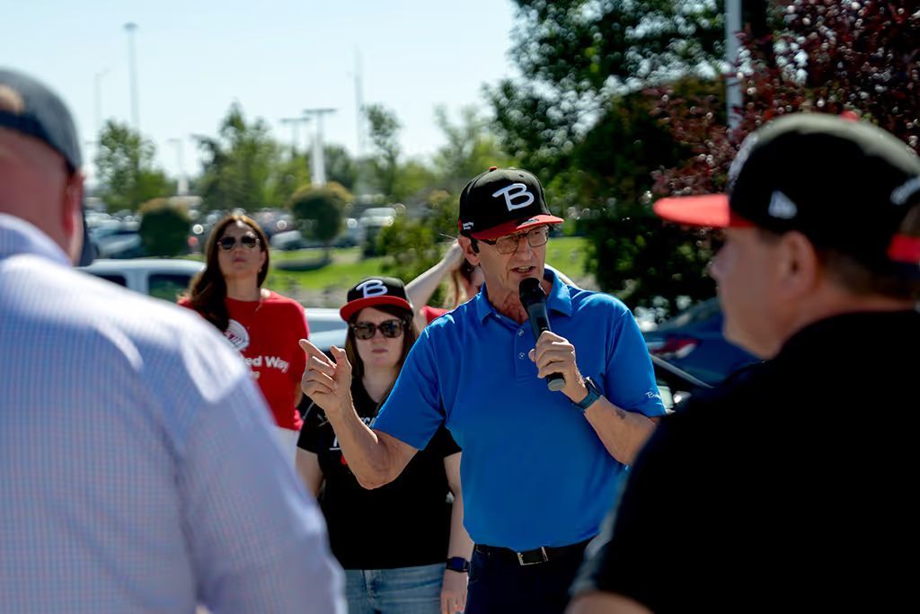 A man in a blue polo shirt and a "B" baseball cap speaking into a microphone during an outdoor event. Several people are visible in the background, including a woman in a red "United Way" shirt.