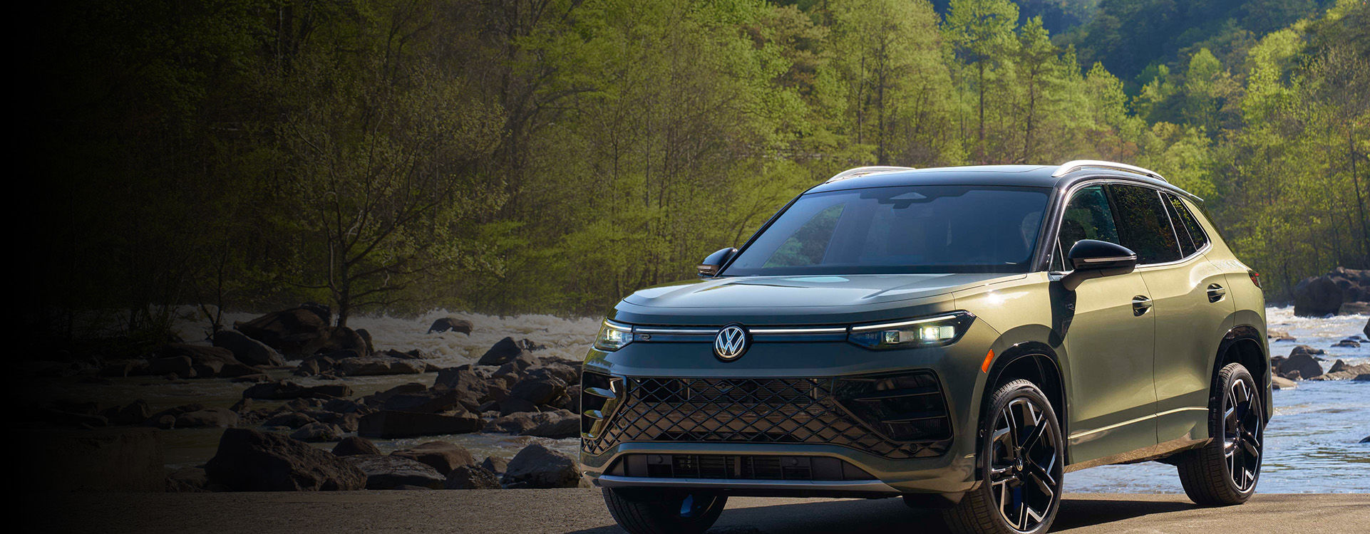 A dark green Volkswagen Taos is parked on a dirt path next to a rocky river with trees and lush greenery in the background, suggesting an outdoor or natural setting.
