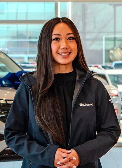 A smiling woman with long dark hair wearing a Birchwood jacket in a car dealership.