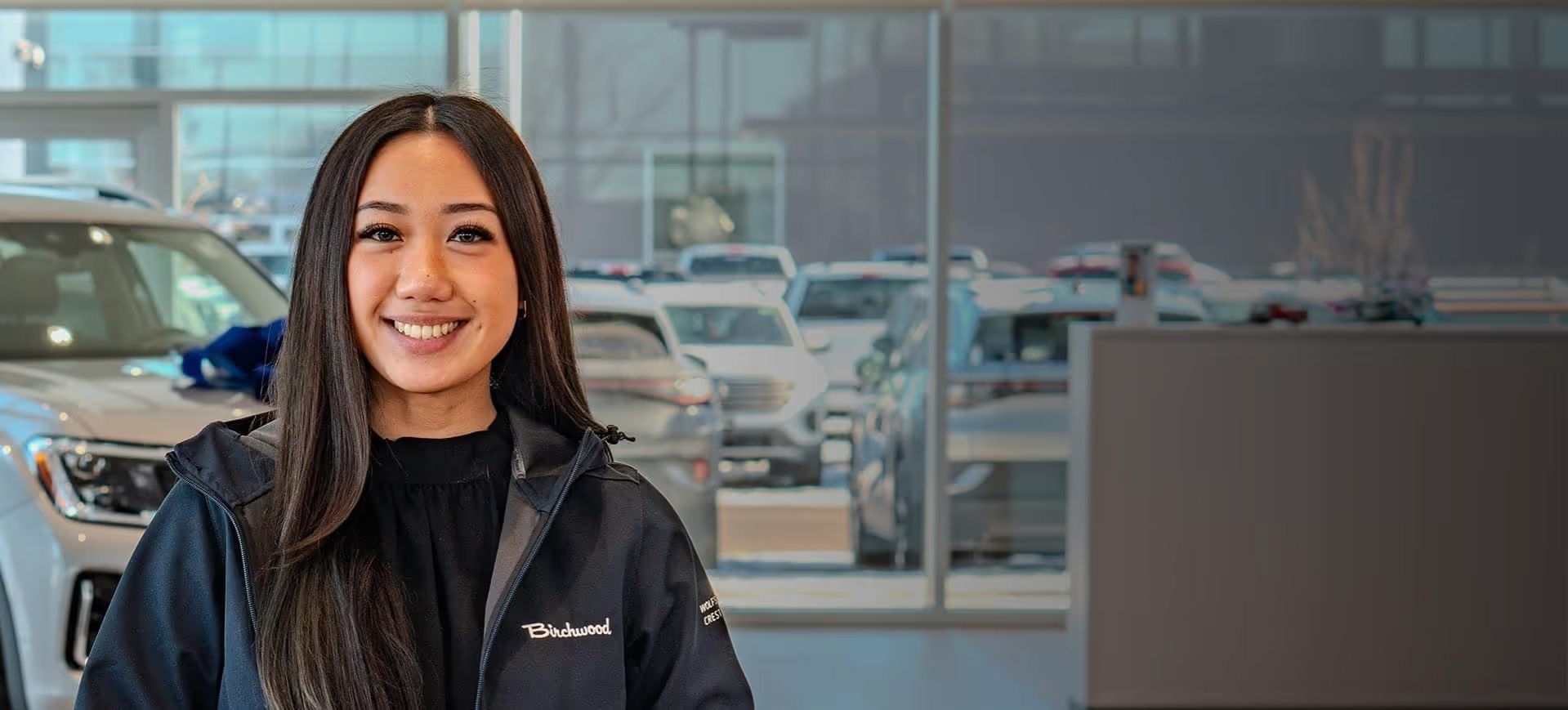 A smiling woman with long dark hair wearing a Birchwood jacket in a car dealership.