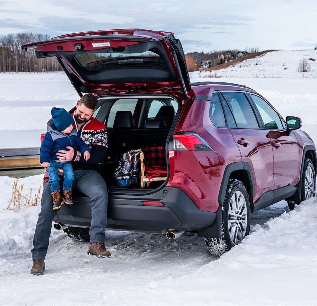 Un père assis sur le rebord du coffre ouvert d'un Toyota RAV4 rouge garé dans la neige, tenant son jeune enfant sur ses genoux. Ils sont tous deux vêtus de vêtements d'hiver.
