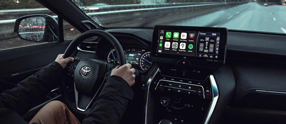 Vue du conducteur à l'intérieur d'un véhicule Toyota moderne (probablement un Venza ou modèle similaire) roulant sur une route mouillée la nuit. Le tableau de bord comporte un grand écran tactile central affichant Apple CarPlay avec diverses icônes d'applications (Téléphone, Messages, Musique, Cartes, etc.).