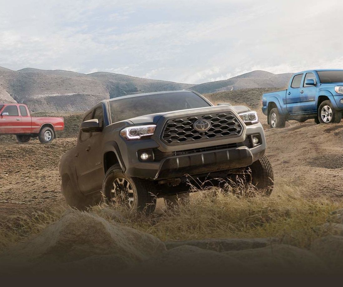 Un Toyota Tacoma gris moderne en conduite hors route, entouré de camions Toyota rouges et bleus plus anciens dans un paysage montagneux et sec.